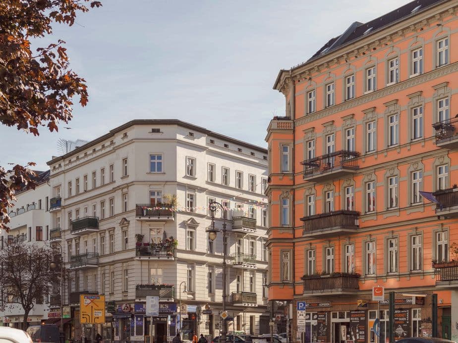 Street view of European-style buildings with ornate balconies, one white and one orange, under a clear sky. Trees and parked cars line the street.