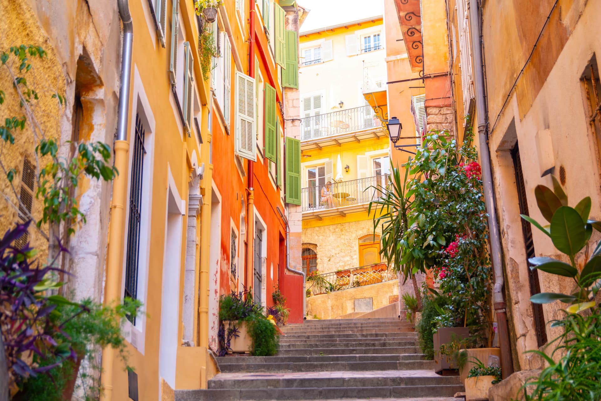 A narrow street with colorful buildings and stairs in Villefranche-sur-Mer, France. The buildings are painted in shades of yellow, orange, and red, with green shutters. Plants are visible along the street.