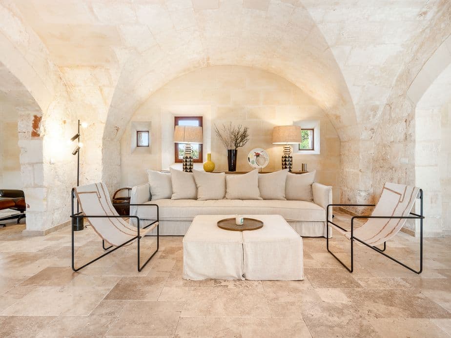 Stylish living room with arched stone ceilings, beige sofa, designer chairs, and modern decor, set in a restored Mediterranean stone house with natural light from small wooden-framed windows.