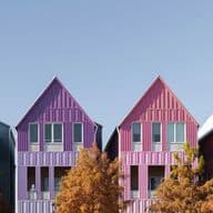 Row of colorful gable-roofed houses in teal, blue, purple, pink, and yellow with autumn trees in front and a clear blue sky.