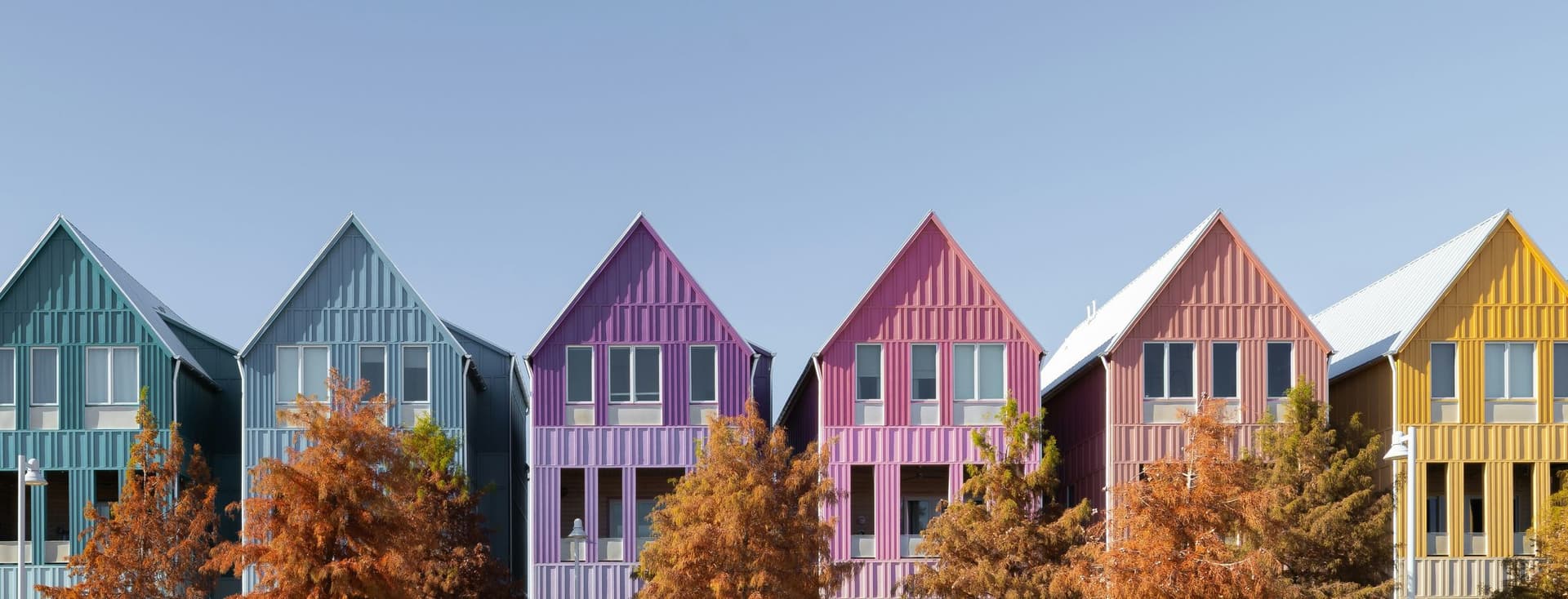 Row of colorful gable-roofed houses in teal, blue, purple, pink, and yellow with autumn trees in front and a clear blue sky.