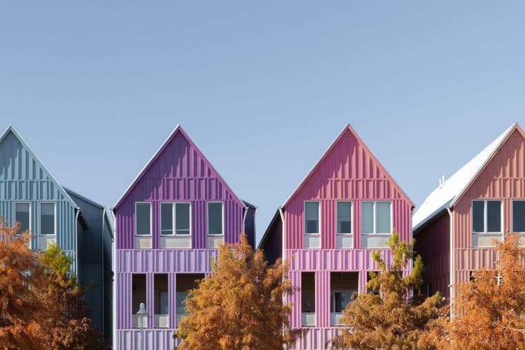 Row of colorful gable-roofed houses in teal, blue, purple, pink, and yellow with autumn trees in front and a clear blue sky.