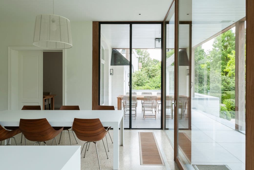 Salle à manger lumineuse avec une table blanche, des chaises en bois et un luminaire moderne. Des portes coulissantes en verre s'ouvrent sur un patio avec une table et des chaises en bois.