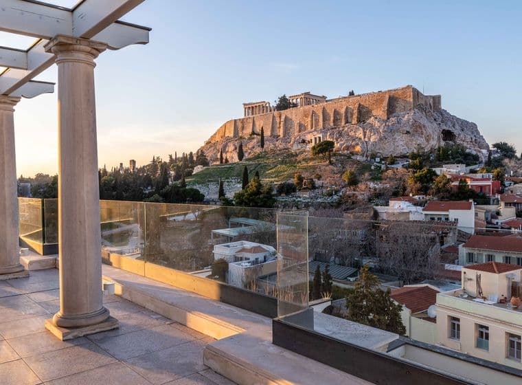 A terrace with columns and glass railings overlooks Athens, featuring the iconic Acropolis and Parthenon atop a rocky hill at sunset. Houses and trees fill the foreground.