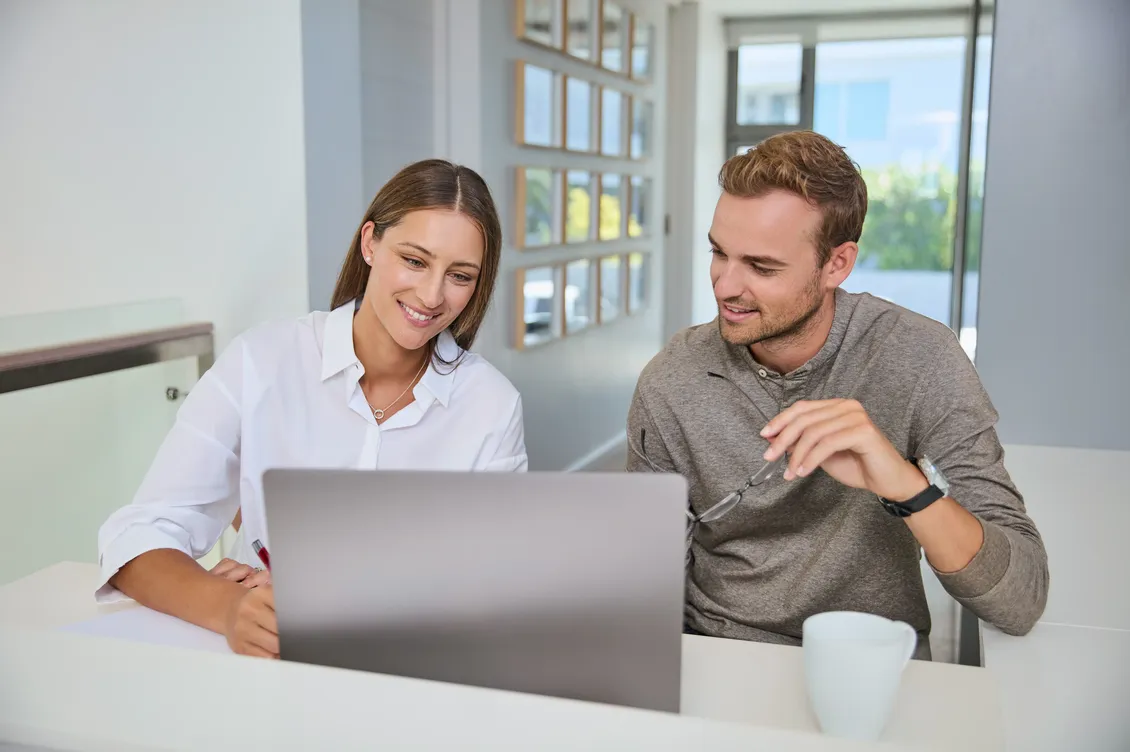 Young couple sitting together at a table, smiling as they look at a laptop, clearly engaged in a consultation or research.