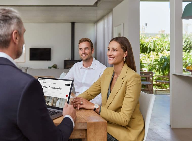 Three people in a modern office setting. A man with a laptop faces a smiling man and woman seated at a wooden table, discussing business.