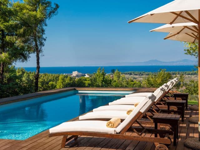 Private swimming pool overlooking the sea and a lush green landscape. Elegant sun loungers line the wooden deck, while panoramic coastal views and surrounding pine trees create a peaceful and exclusive atmosphere.