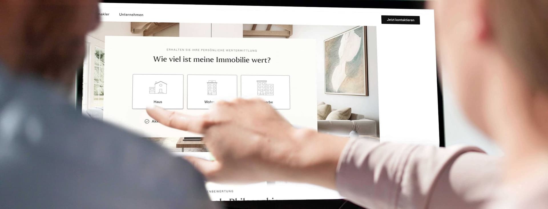Two people pointing at a laptop showing a German real-estate valuation page reading "Wie viel ist meine Immobilie wert?"