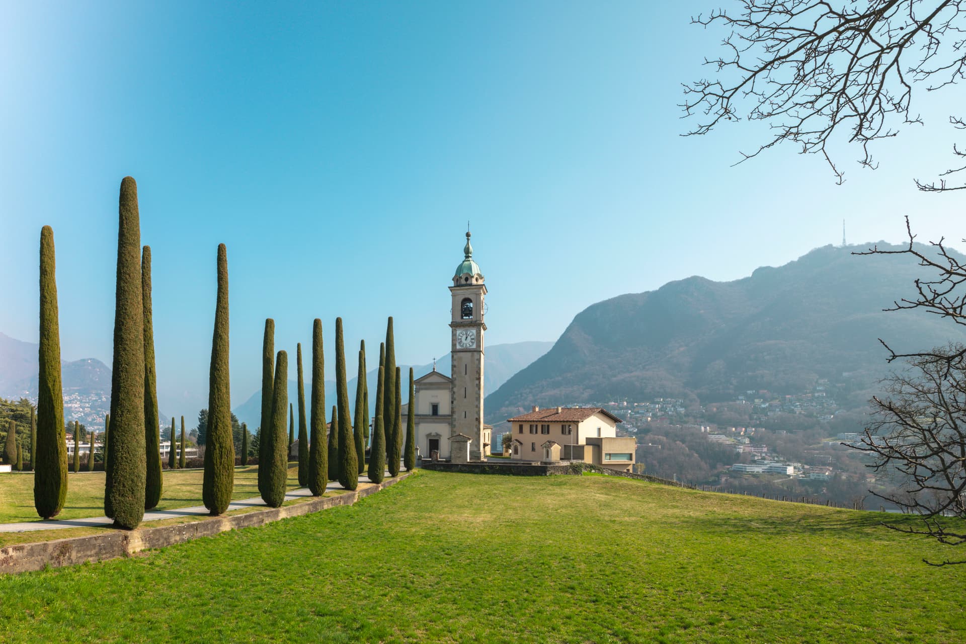 Row of tall cypress trees and a small church bell tower with clock on a grassy hillside against mountains and clear blue sky.