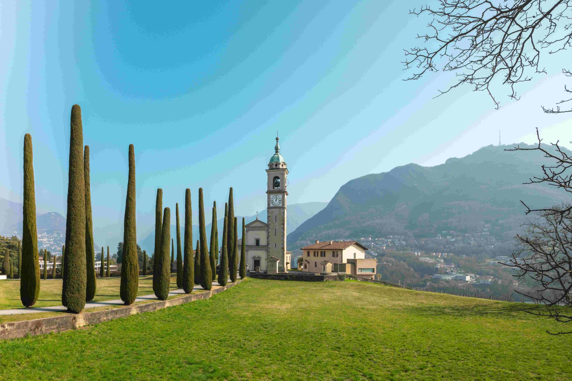 Tall cypress trees leading to a church bell tower with clock, grassy hill and distant mountains under a clear blue sky.