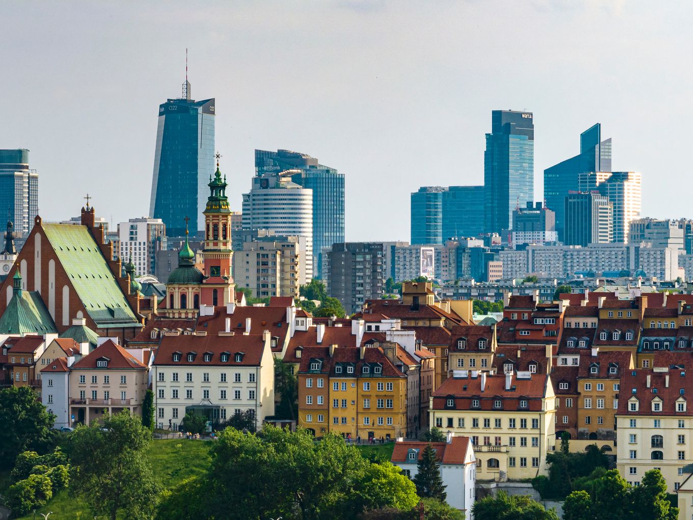 Panoramic view of Warsaw, showing a mix of historic buildings and modern skyscrapers under a clear sky, with trees in the foreground.