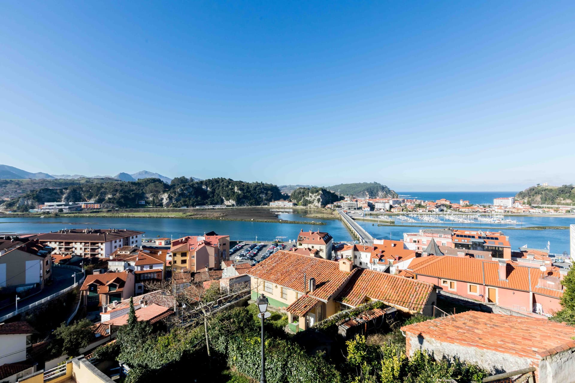 Vista aèria de Ribadesella, Espanya, amb edificis de teulades vermelles, un riu, un pont i muntanyes sota un cel blau clar.