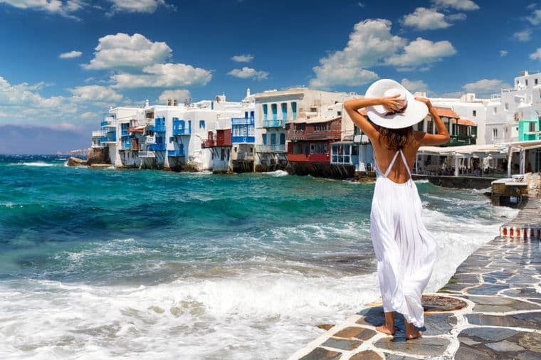 A woman in a white dress and hat looks out at the sea in Mykonos, Greece. White buildings with blue accents line the shore.