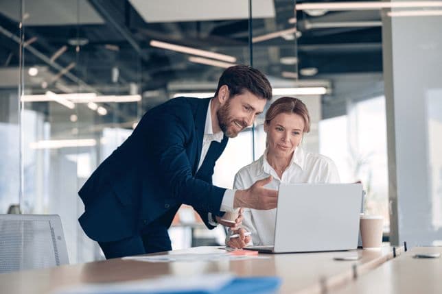 A man in a suit points at a laptop screen as a woman in a white shirt looks on in an office.