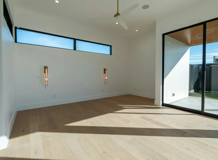 Bright, modern empty room with light wood flooring, large glass sliding doors to an outdoor patio, a ceiling fan, and a high horizontal window.