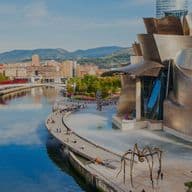 Blick auf das Guggenheim Museum Bilbao, Spanien, mit dem Nervión-Fluss, einer großen Spinnenskulptur und Menschen, die herumlaufen.