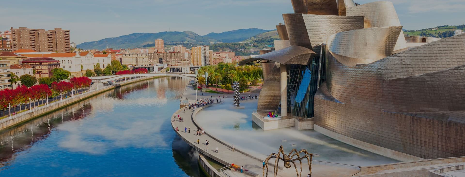 Vista del Museu Guggenheim Bilbao, Espanya, amb el riu Nervión, una gran escultura d'aranya i gent passejant al voltant.