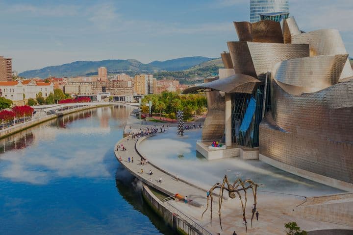 Blick auf das Guggenheim Museum Bilbao, Spanien, mit dem Nervión-Fluss, einer großen Spinnenskulptur und Menschen, die herumlaufen.