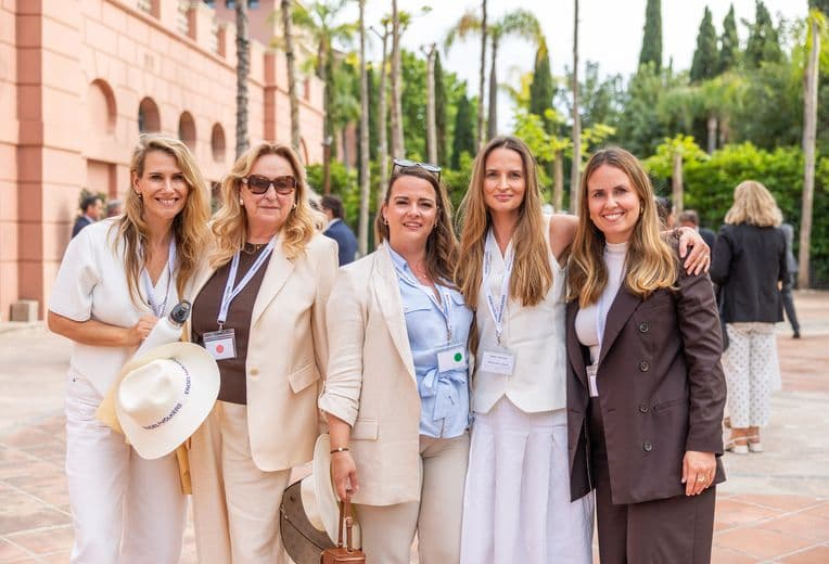 Five women in light outfits with lanyards pose together outdoors, in front of a pink arched building and palm trees.