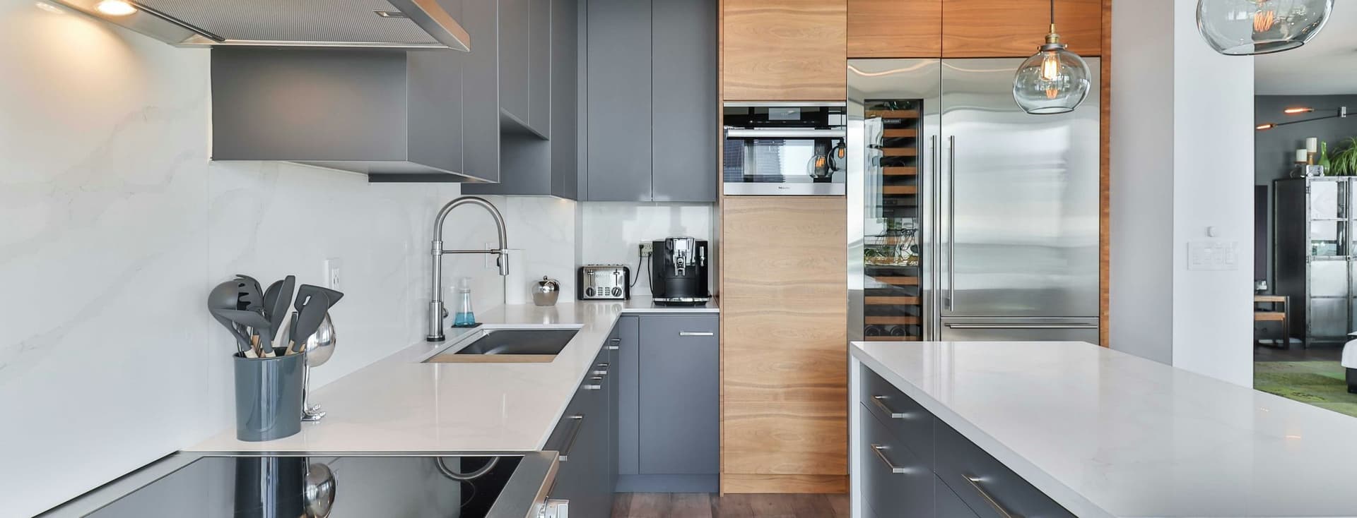Sleek kitchen with gray cabinets, wooden pantry accents, stainless steel appliances, a white island, and two glass pendant lights.