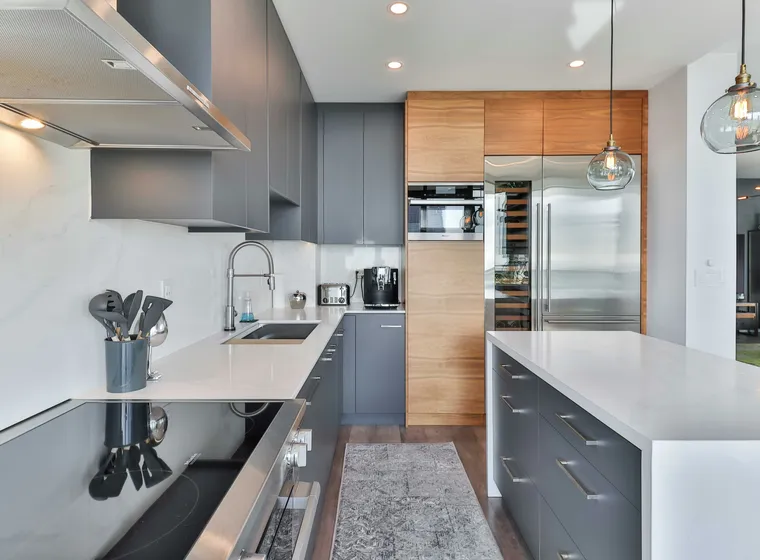 Sleek kitchen with gray cabinets, wooden pantry accents, stainless steel appliances, a white island, and two glass pendant lights.