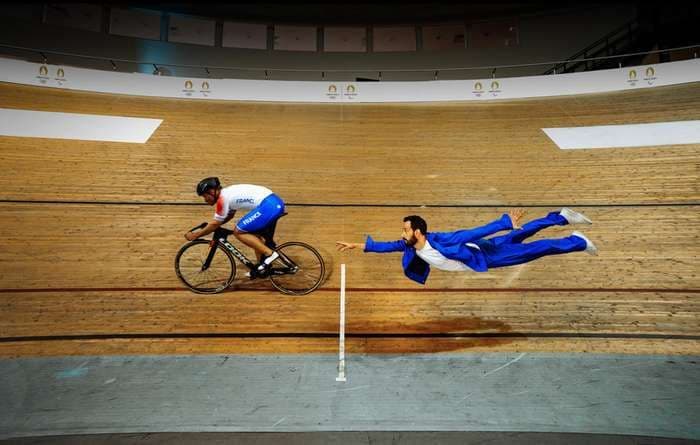 Indoor cycling track with a cyclist and a man in a blue suit appearing to fly. The cyclist wears a white and blue jersey.