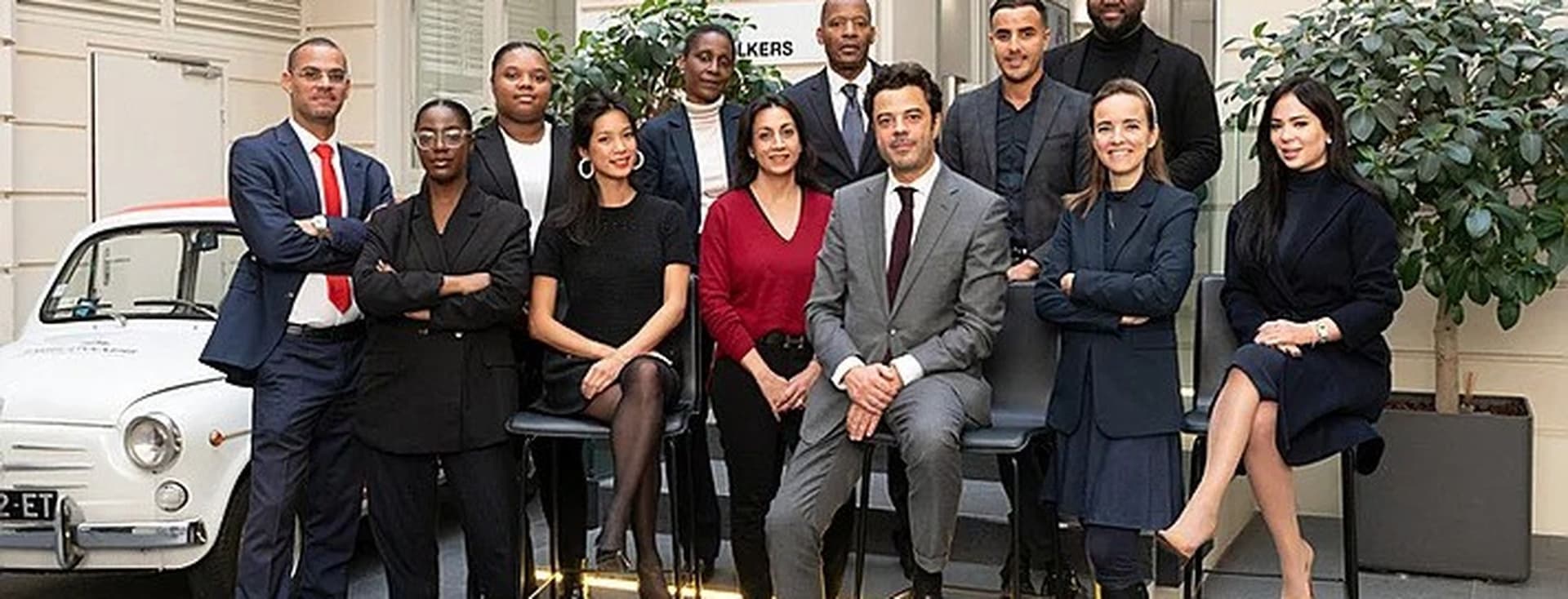 A diverse real estate team poses in suits and dresses in front of a white vintage car.