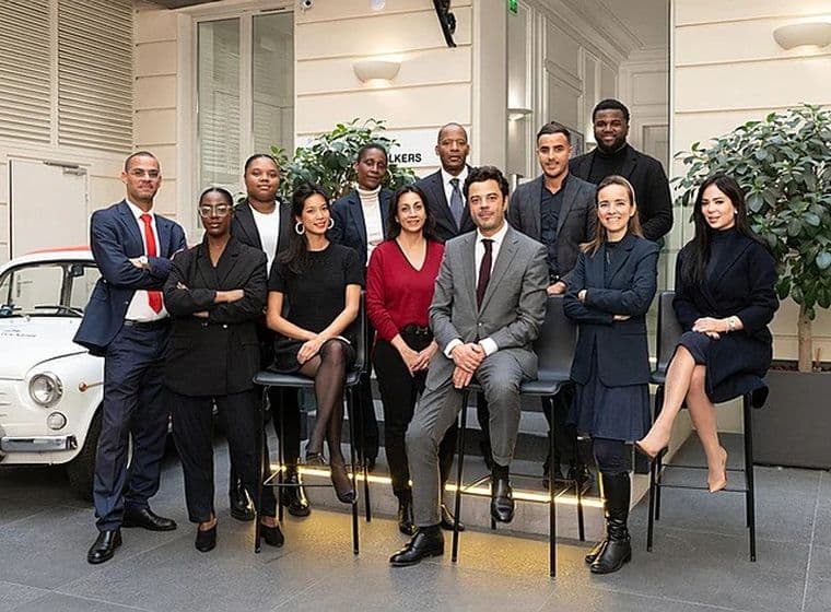 Une équipe immobilière diversifiée pose en costumes et robes devant une voiture ancienne blanche.