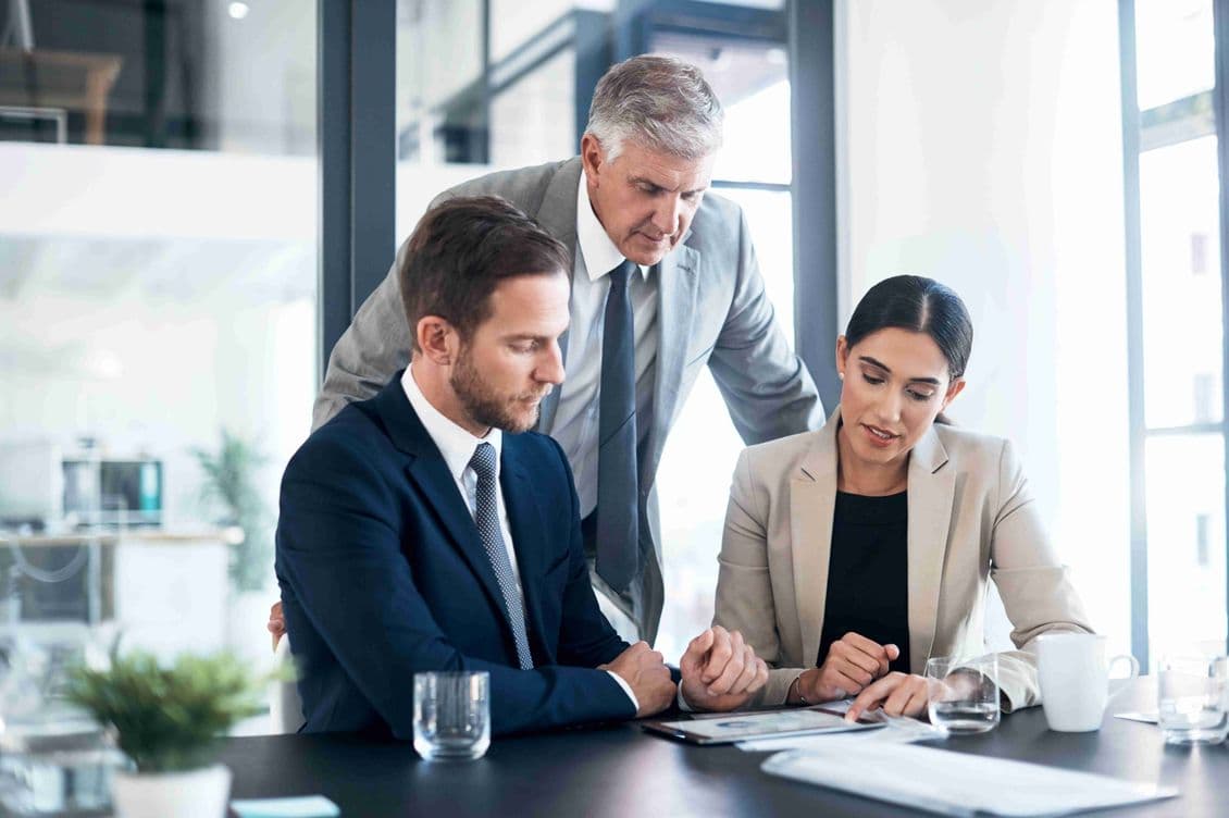 Three business people in suits review data on a tablet at a table in a bright office.