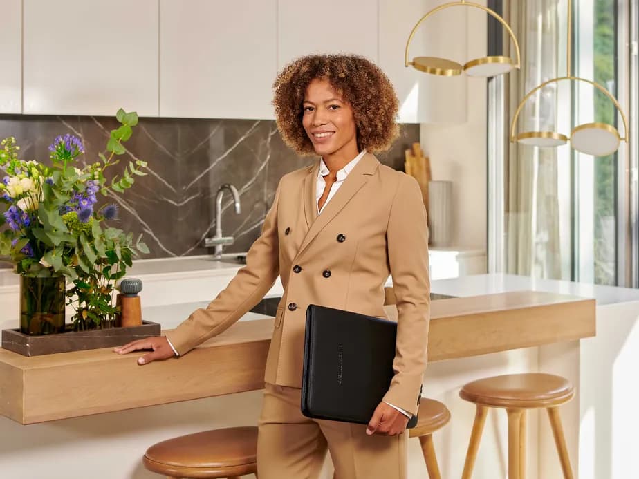 A smiling woman wearing a beige suit stands beside the island in a modern kitchen, with a laptop, flowers and gold pendant lights.