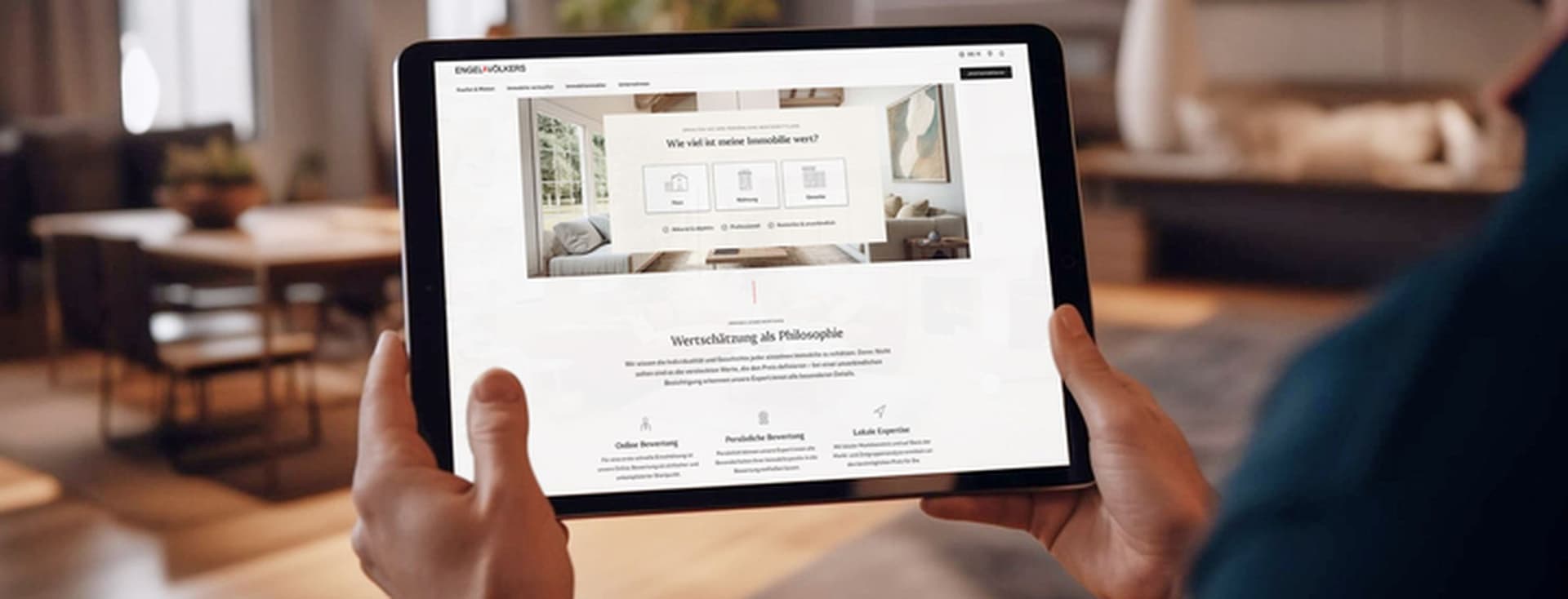 Um homem segura um tablet exibindo o site de imóveis da Engel & Völkers em uma sala de estar moderna.