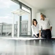 Two people in white shirts review blueprints on a table near a large window in a bright, modern office.