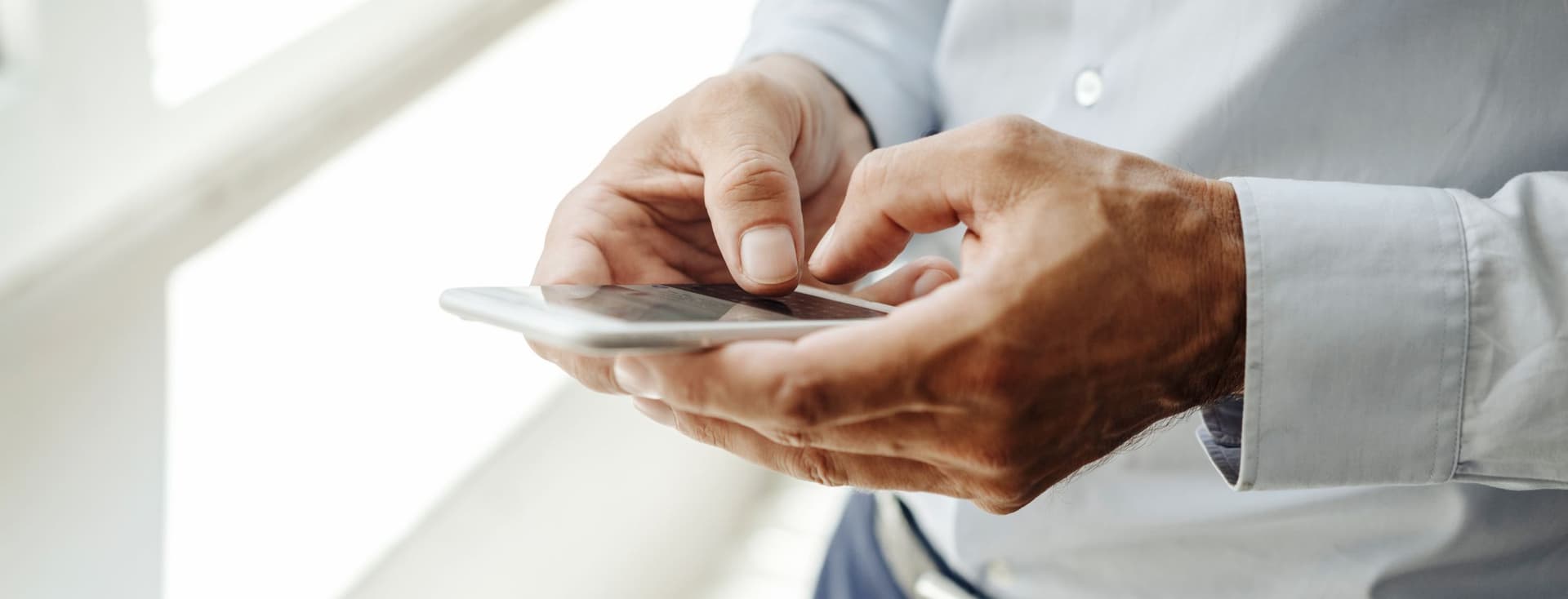 Close-up of business professional's hands in white shirt sleeve using smartphone, photographed against bright white background in modern office environment