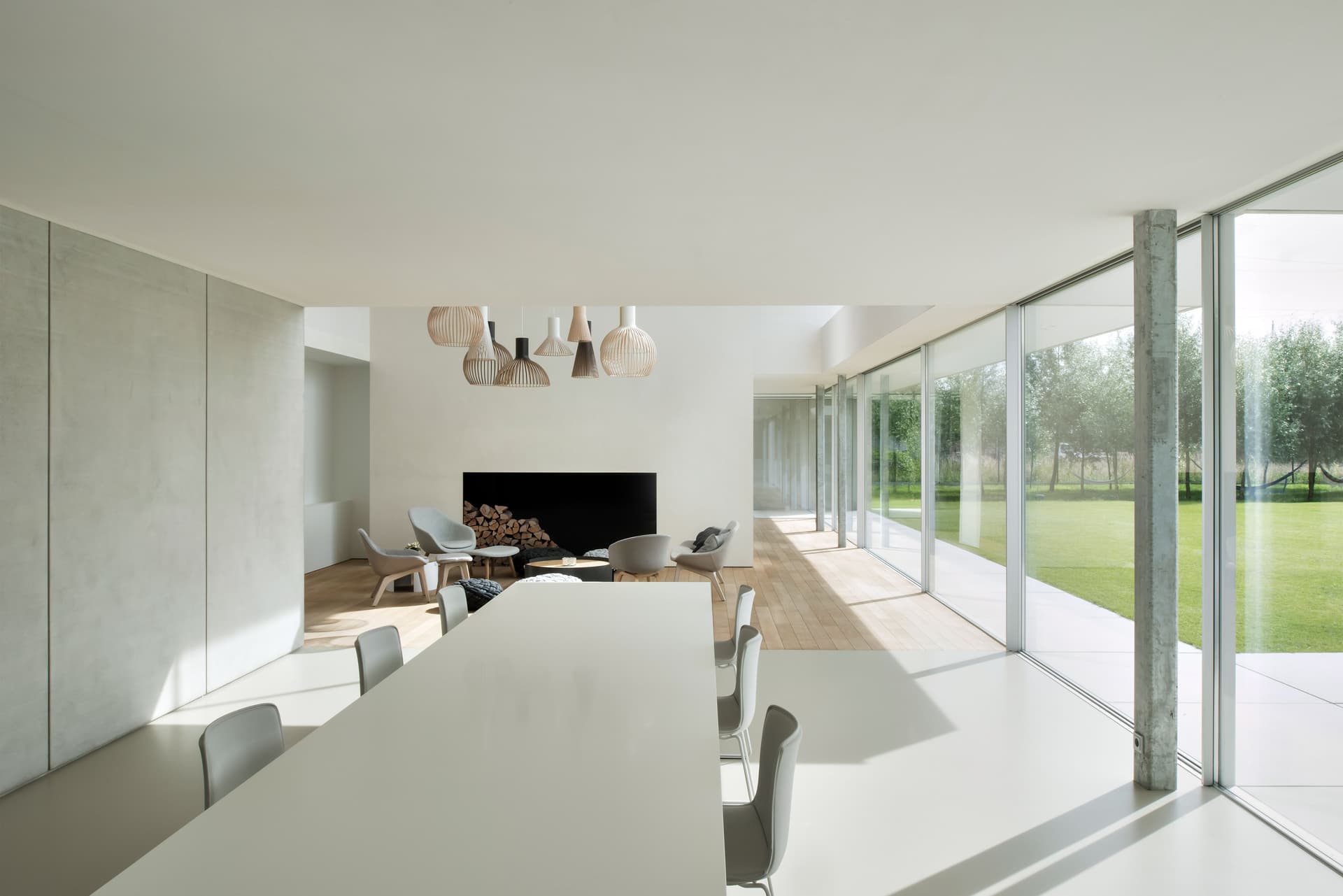 Helles, modernes Interieur mit weißen Wänden, großen Fenstern und einem Esstisch. Ein Kamin und eine Sitzecke sind im Hintergrund sichtbar.