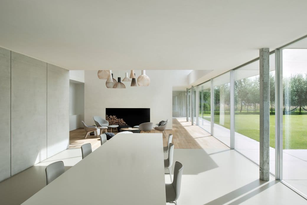 Bright, modern interior with white walls, large windows, and a dining table. A fireplace and seating area are visible in the background.