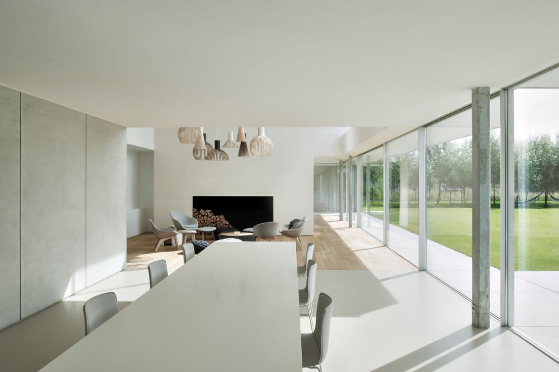 Bright, modern interior with white walls, large windows, and a dining table. A fireplace and seating area are visible in the background.