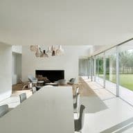 Bright, modern interior with white walls, large windows, and a dining table. A fireplace and seating area are visible in the background.