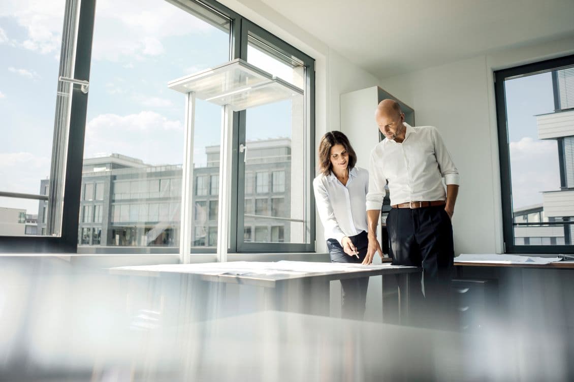 Two people in white shirts review plans on a table in a bright office with a large window.