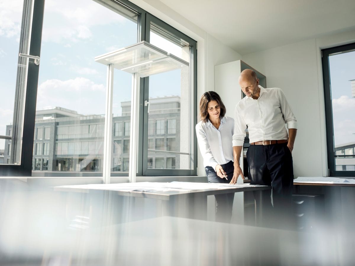 Two people in white shirts review plans on a table in a bright office with a large window.