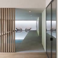 Minimalist indoor pool seen through a corridor; vertical wooden slats on the left, glass wall on the right, and two lounge chairs by the pool.