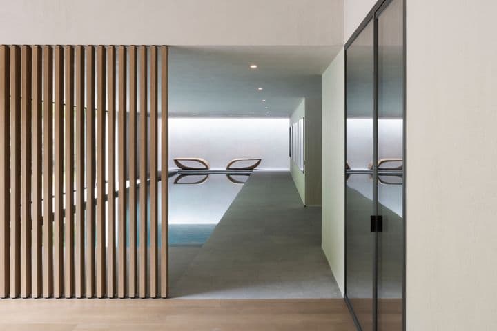 Minimalist indoor pool seen through a corridor; vertical wooden slats on the left, glass wall on the right, and two lounge chairs by the pool.