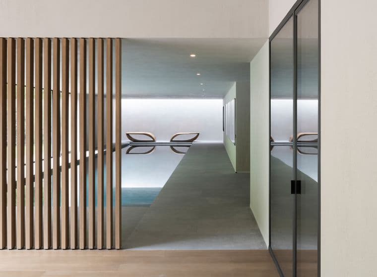 Minimalist indoor pool seen through a corridor; vertical wooden slats on the left, glass wall on the right, and two lounge chairs by the pool.