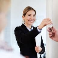 A smiling female real estate agent in a black blazer and white shirt extending her arm to hand over documents, captured in a bright professional office setting. Her expression conveys warmth and professionalism.