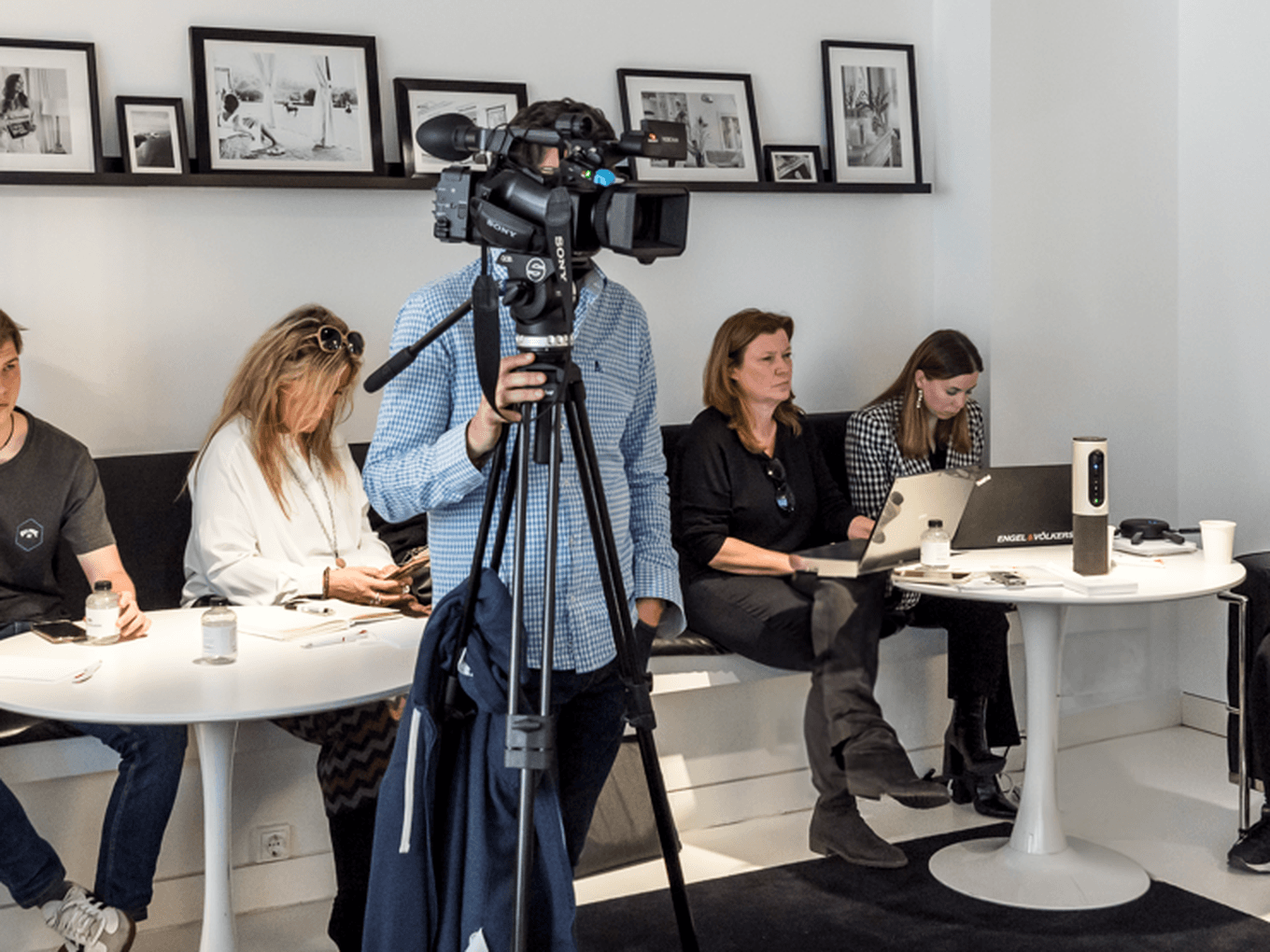 Uma pessoa filma um painel de pessoas sentadas em mesas brancas numa sala branca com fotos a preto e branco numa prateleira.
