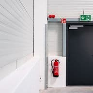 Industrieller Flur mit einer schwarzen Notausgangstür, einem grünen Ausgangsschild darüber und einem roten Feuerlöscher an der Wand. Weiße Wände und Innenausstattung im Industriestil.