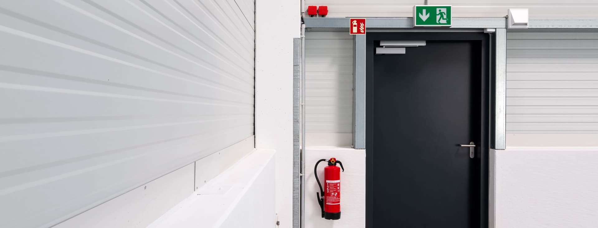 Industrial hallway with a black emergency exit door, green exit sign above, and red fire extinguisher mounted on the wall. White walls and industrial-style interior.