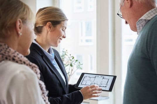 A female real estate agent in a black blazer showing floor plans on a tablet to two clients - a woman in a light coat with a patterned scarf and a man wearing glasses - in a bright, window-lit office setting.