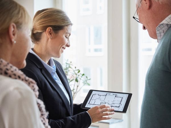 A real estate agent shows a house plan on a tablet to an elderly couple. The agent is smiling.