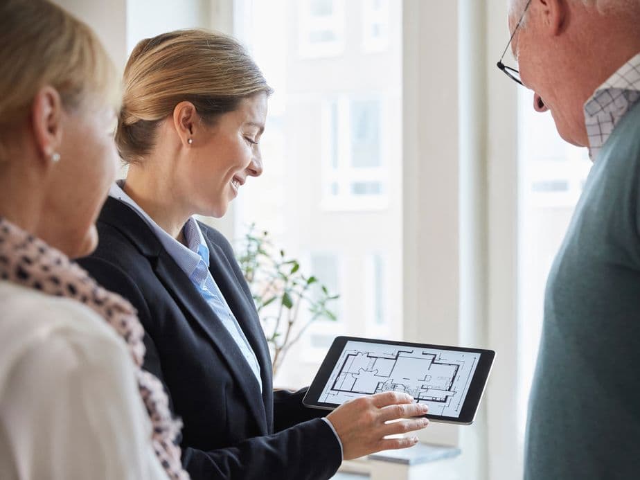 A female real estate agent in a black blazer showing floor plans on a tablet to two clients - a woman in a light coat with a patterned scarf and a man wearing glasses - in a bright, window-lit office setting.