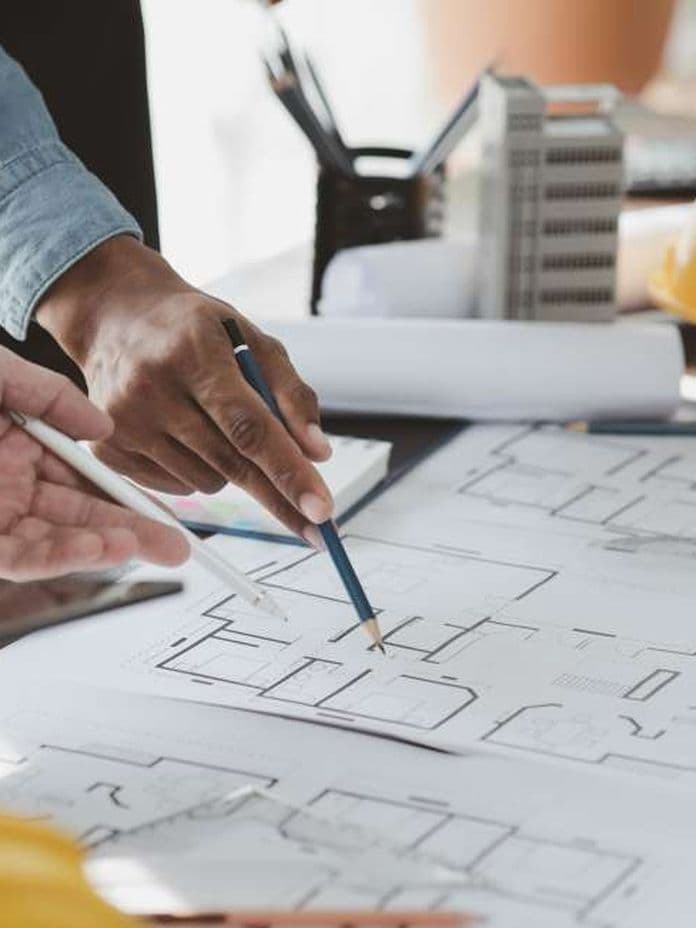 People discussing architectural blueprints on a table, with a yellow hard hat and building model in the background.